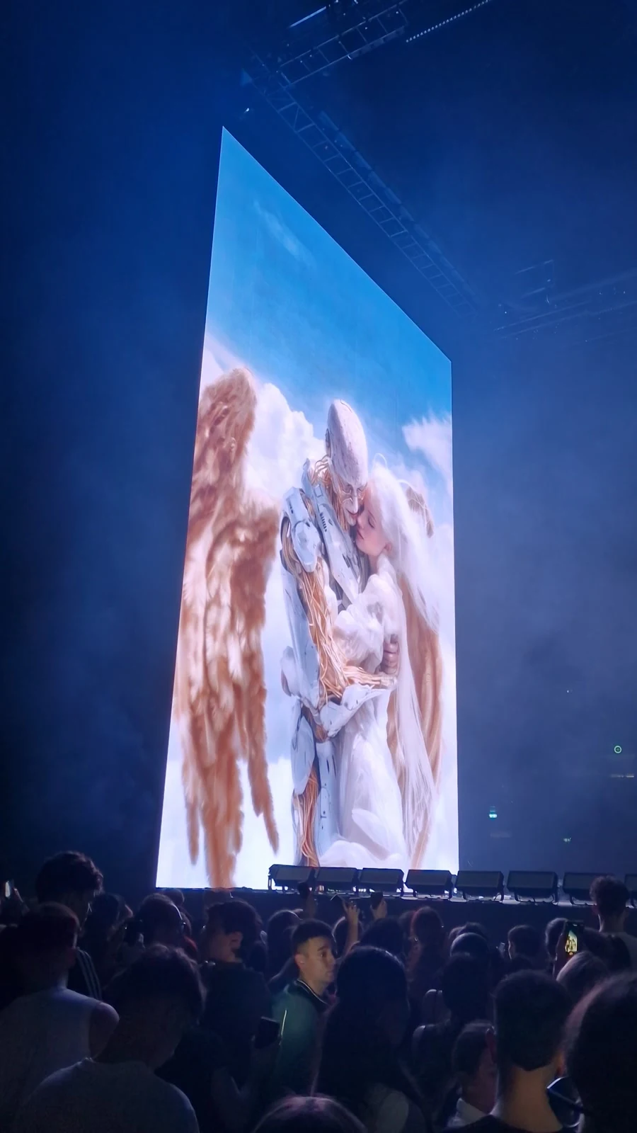 A tall stage screen showing winged figures, with the concert audience in the foreground.