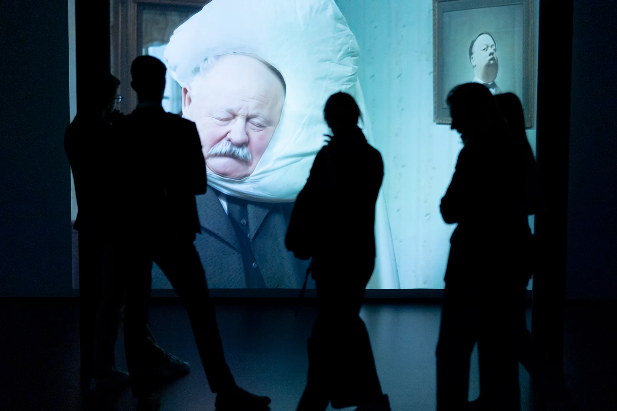Visitors silhouetted in front of a large projection of a suited figure with a white form surrounding their head at MEET.